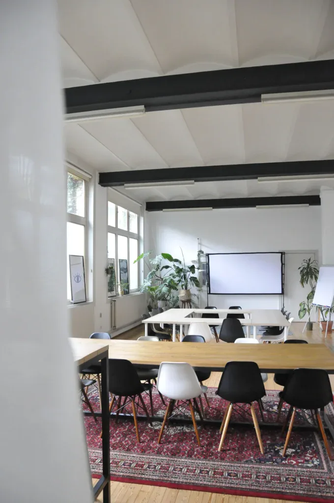 Ruime vergaderruimte met U-opstelling, projectiescherm en veel daglicht, geschikt voor presentaties en interactieve sessies.