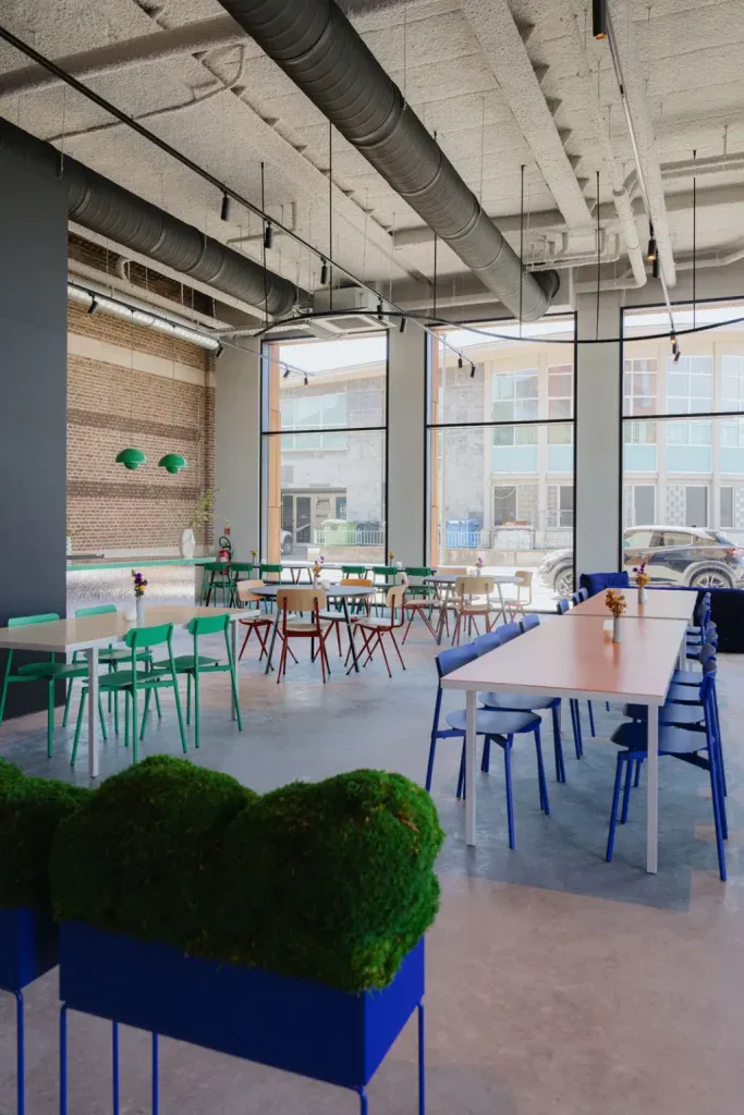 Lichtrijke zaal met industriële architectuur, lange tafels en kleurrijke stoelen, geschikt voor workshops en zakelijke events
